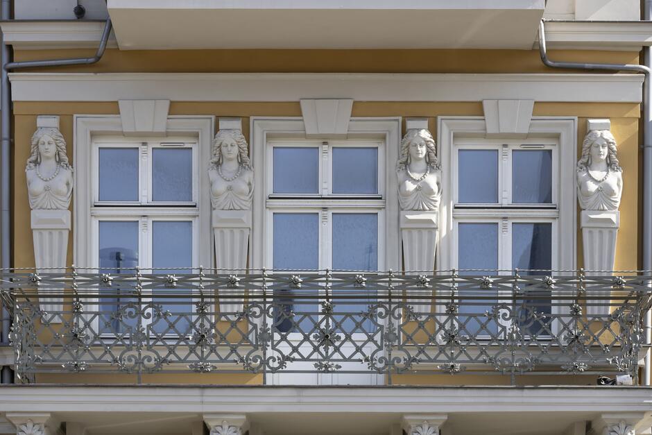 Zdjęcie przedstawia fragment fasady klasycznego budynku, skupiając się na eleganckim balkonie z ozdobną, metalową balustradą. Nad balustradą widzimy szereg białych okien z niebieskimi firankami, które są ujęte między klasycznymi, rzeźbionymi postaciami kariatyd, wspierającymi architektoniczne nadproża. Kariatydy mają upięte włosy i są ubrane w stroje stylizowane na antyczne, co dodaje elewacji wyrafinowanego charakteru. Kolory fasady są stonowane, z dominującą ciepłą żółcią i bielą, które ładnie kontrastują z niebieskimi elementami okien. Budynek reprezentuje klasycystyczny styl architektoniczny, a jego zdobienia i detale świadczą o zamiłowaniu do estetyki i form zaczerpniętych z antyku