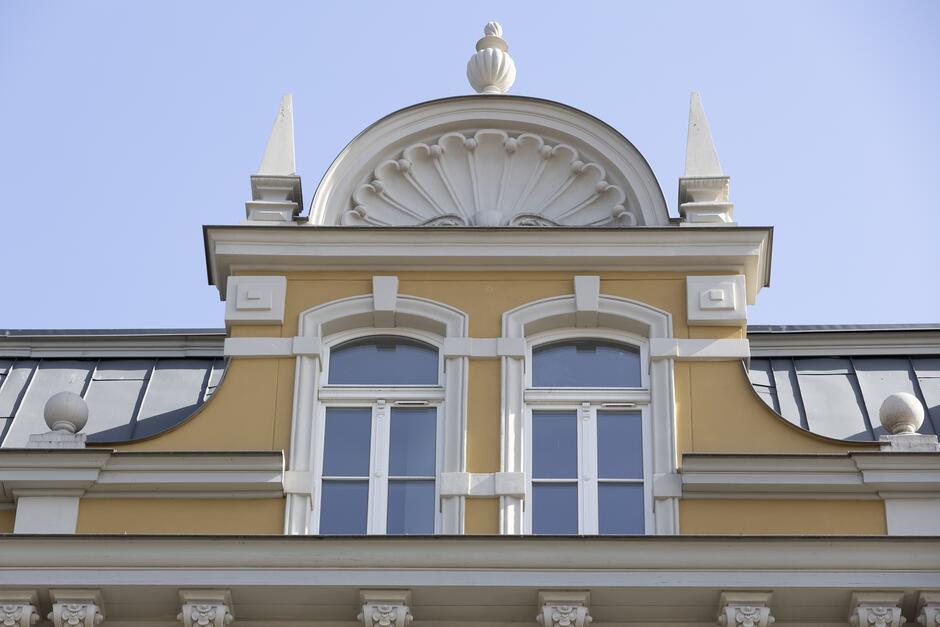 Zdjęcie pokazuje górną część klasycznej fasady budynku. Centralnym elementem jest półkolisty fronton zwieńczony dekoracyjnym motywem muszli i kulą na szczycie. Fronton jest wsparty przez białe pilastry, które flankują trzy okna z łukowatymi górnymi częściami, podkreślone białymi opaskami. Całość umieszczona jest na tle żółtej elewacji. Powyżej okien widać detale gzymsu z motywami klasycznymi, a na końcach wystające, ozdobne elementy w kształcie ostrosłupów. Struktura dachu to mansardowy, odcinający się ciemnymi panelami, dodający elegancji całej konstrukcji. Ten fragment budynku odzwierciedla klasyczne, europejskie podejście do architektury, gdzie każdy detal jest starannie zaprojektowany i skomponowany, tworząc harmonijny i wyrafinowany obraz.