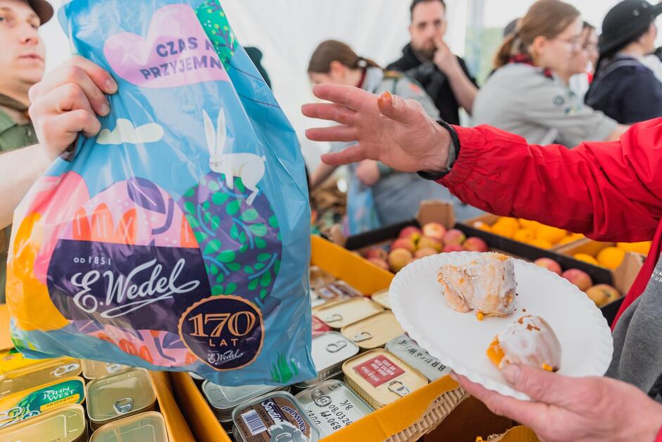 Zdjęcie ukazuje wydawanie żywności podczas jakiegoś wydarzenia. Na pierwszym planie jest osoba w czerwonym rękawie, która trzyma papierowy talerzyk z kawałkami ciasta lub deseru. W tle widoczny jest wolontariusz trzymający dużą, kolorową torbę z napisem CZAS PRZYJEMNY  i logo  E.Wedel 170 lat , co sugeruje, że jest to torba z produktami tej znanej polskiej firmy cukierniczej, świętującej swoje jubileum. W tle, w centralnej części zdjęcia, widać inne osoby zajęte organizacją wydarzenia. Po prawej stronie jest karton z puszkami, który może zawierać inne artykuły spożywcze. Całość sprawia wrażenie aktywnego udziału w jakimś wydarzeniu charytatywnym lub społecznym, gdzie ludzie są obdarowywani żywnością.