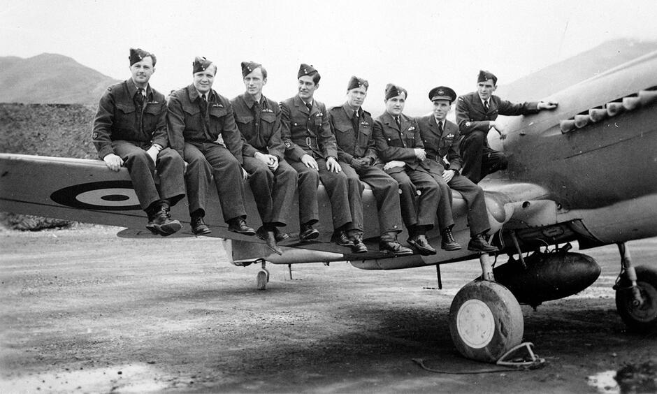 Czarno-białe zdjęcie. Mężczyźni siedzą na skrzydle samolotu wojskowego