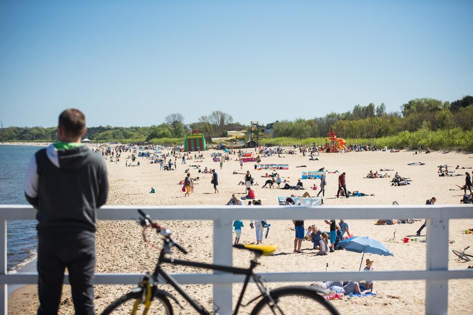 na zdjęciu chłopak z rowerem, stoi na promenadzie, w tle plaża i mnóstwo plażowiczów