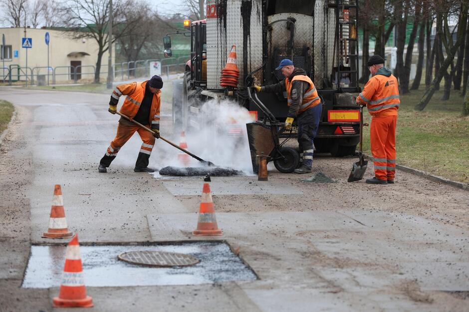 Trzech mężczyzn w roboczych, pomarańczowych kombinezonach naprawia nawierzchnię ulicy, wylewając asfalt.