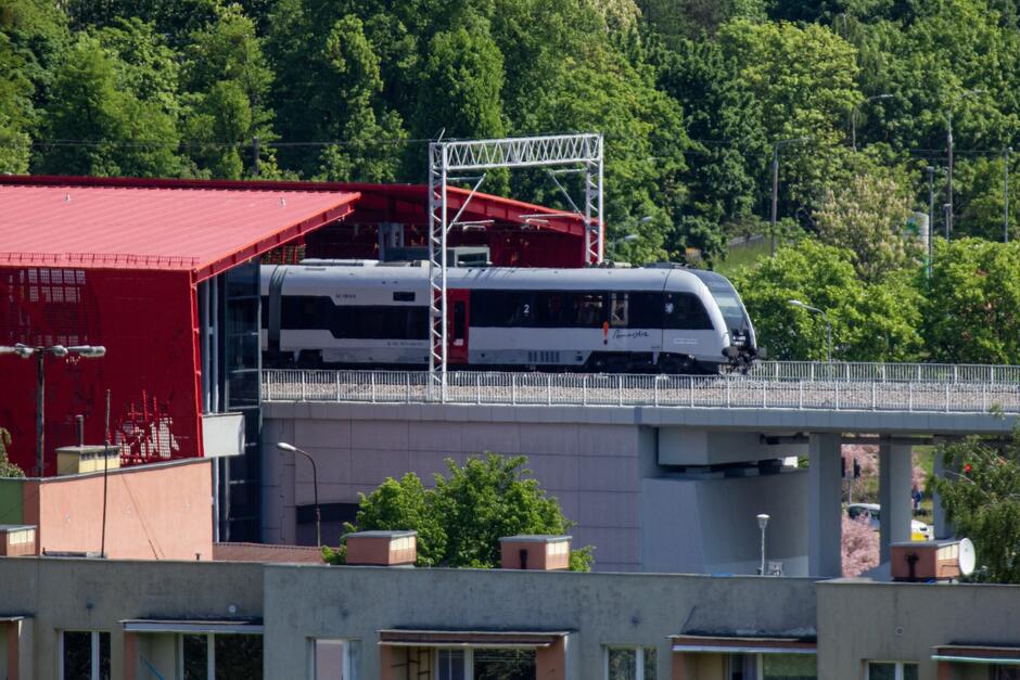 zdjęcie przedstawia pociąg pasażerski przejeżdżający przez most kolejowy. Pociąg ma nowoczesny wygląd, z zaokrąglonym przodem, charakterystyczny dla współczesnych szybkich składów. Kolorystyka składa się głównie z bieli z akcentami kolorów, w tym czerwonego dachu widocznego budynku w tle, który kontrastuje z zielenią drzew w oddali. Na pierwszym planie widzimy budynek, być może mieszkalny, z balkonami. Scena jest nasłoneczniona, co wskazuje na pogodny dzień.