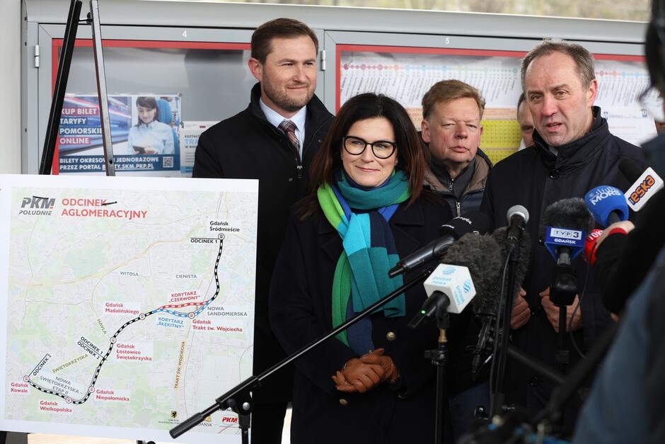 Cztery osoby przy tablicy informującej o przebiegu nowej linii kolejowej PKM. Tablica znajduje się po lewej, osoby stoją centralnie
