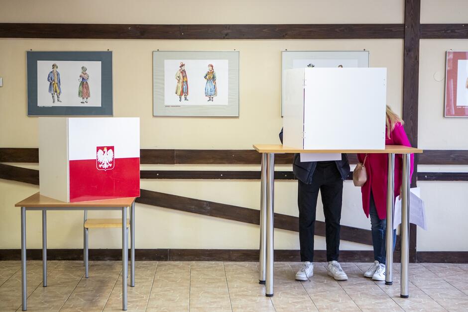 Na zdjęciu widzimy wnętrze lokalu wyborczego, gdzie odbywa się proces głosowania. Po lewej stronie ustawiono jedną z tradycyjnych czerwono-białych urn wyborczych z godłem Polski – białym orłem na czerwonym tle. Po prawej stronie znajduje się osoba w trakcie głosowania, stojąca za mobilną kabiną wyborczą, która zapewnia prywatność. Osoba ma na sobie różową górną część ubrania i ciemne spodnie; obok niej widać białą kartę do głosowania. W tle na ścianie zawieszono obrazy przedstawiające postacie w tradycyjnych polskich strojach ludowych, co nadaje pomieszczeniu lokalny charakter. Pomieszczenie jest jasne i wydaje się być publiczną przestrzenią, być może w szkole lub urzędzie.