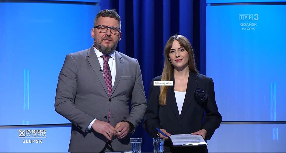 Na zdjęciu widzimy dwie osoby stojące obok siebie w studiu telewizyjnym. Po lewej stronie jest mężczyzna w szarym garniturze, białej koszuli i czerwonym krawacie w drobny wzór, z okularami. Po prawej stronie stoi kobieta w eleganckim, czarnym garniturze z bluzką o jasnym kolorze, jej włosy są długie i jasnobrązowe. Oboje wyglądają na prezenterów lub moderatorów, kobieta trzyma w rękach dokumenty. Tło jest jednolicie niebieskie z wyraźnym logo TVP3 Gdańsk w prawym górnym rogu. W lewym dolnym rogu znów pojawia się napis POMORZE WYBIERA - SŁUPSK , sugerujący tematyczny kontekst programu. Na środku u dołu obrazu jest mały, nieaktywny element interfejsu użytkownika z napisem  Odtwarzaj wideo , co wskazuje, że zdjęcie może pochodzić z platformy streamingowej lub z nagrania wideo