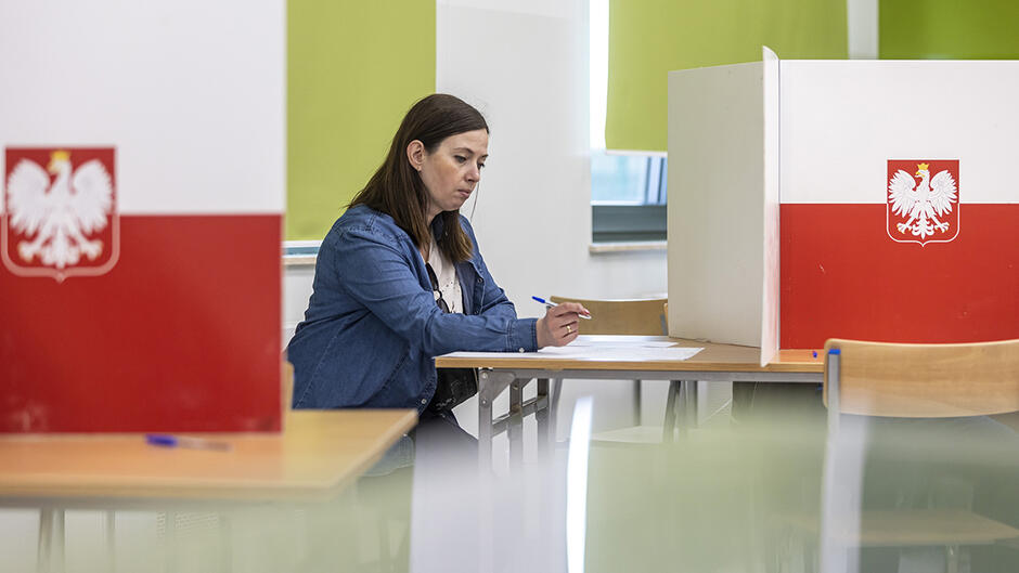 Kobieta siedzi przy stoliku. Po bokach inne stoliki z ustawionymi kartami w biało-czerwonych barwach z orłem