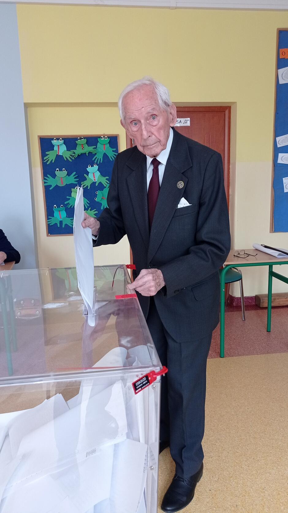 Starszy mężczyzna w garniturze stoi przy urnie wyborczej, wkłada do niej kartki 