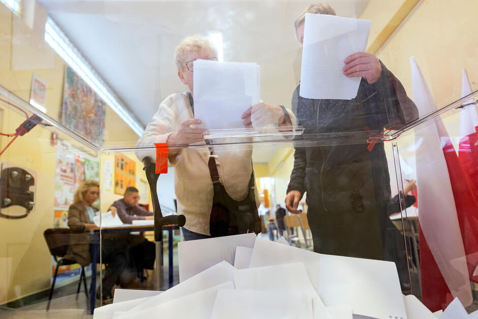 Na pierwszym planie urna wyborcza z kartami (widok z dołu), nad nią dwie osoby z kartami (karty zasłaniają głowy). W tle stoły z osobami siedzącymi za nimi 