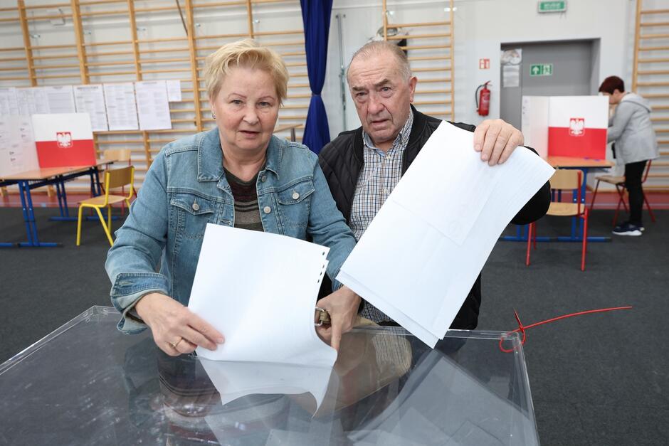 Dwoje starszych ludzi (kobieta i mężczyzna) stoją z kartami przed urną wyborczą