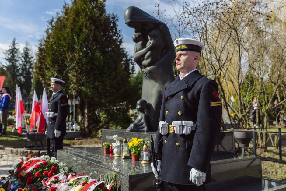 Na zdjęciu widzimy mężczyznę w mundurze marynarki wojennej, z charakterystycznymi białymi rękawiczkami i czapką, stoi na tle wspomnianego w tekście pomnika. W tle widoczne są flagi i wieńce. Otoczenie jest zielone i nasłonecznione.