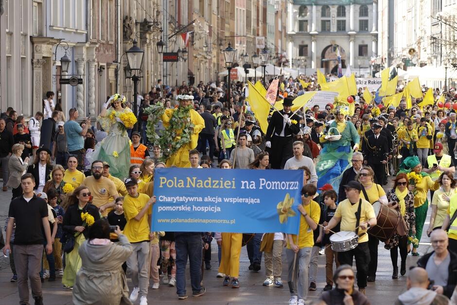Z lewej strony fragmenty kamienic w centrum parada osób w różnym wieku, kobiet i mężczyzn, dzieci, idą w żółtych koszulkach, na przodzie niosą transparent Pola Nadziei na Pomorzu
