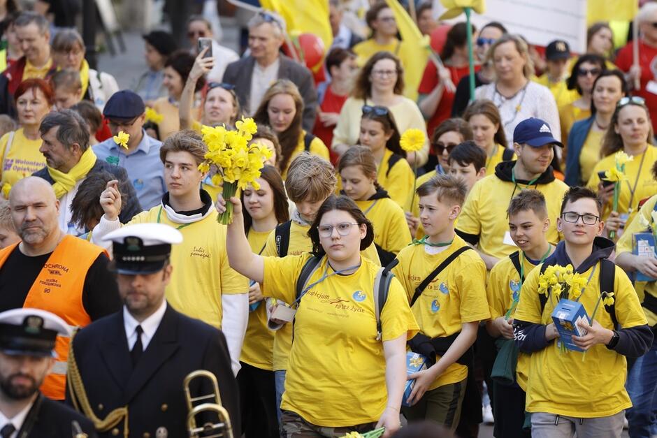 Tłum dziewcząt i chłopców w żółtych koszulkach, niektórzy trzymają w dłoniach bukiety żółtych kwiatów. Z lewej strony idzie mężczyzna w mundurze