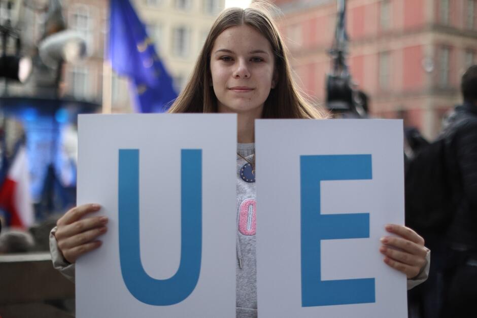 Na zdjęciu widzimy młodą kobietę, która trzyma dwa białe plakaty z literami U  i  E  w kolorze niebieskim, które razem tworzą skrót  UE , co jest powszechnie używanym skrótem dla Unii Europejskiej. Kobieta ma długie, proste brązowe włosy i jest ubrana w szary top. Stoi na tle zatłoczonego miejsca, prawdopodobnie na jakimś zgromadzeniu lub demonstracji, co sugerują nieostre tło i widoczne flagi. Na jej twarzy maluje się delikatny uśmiech.