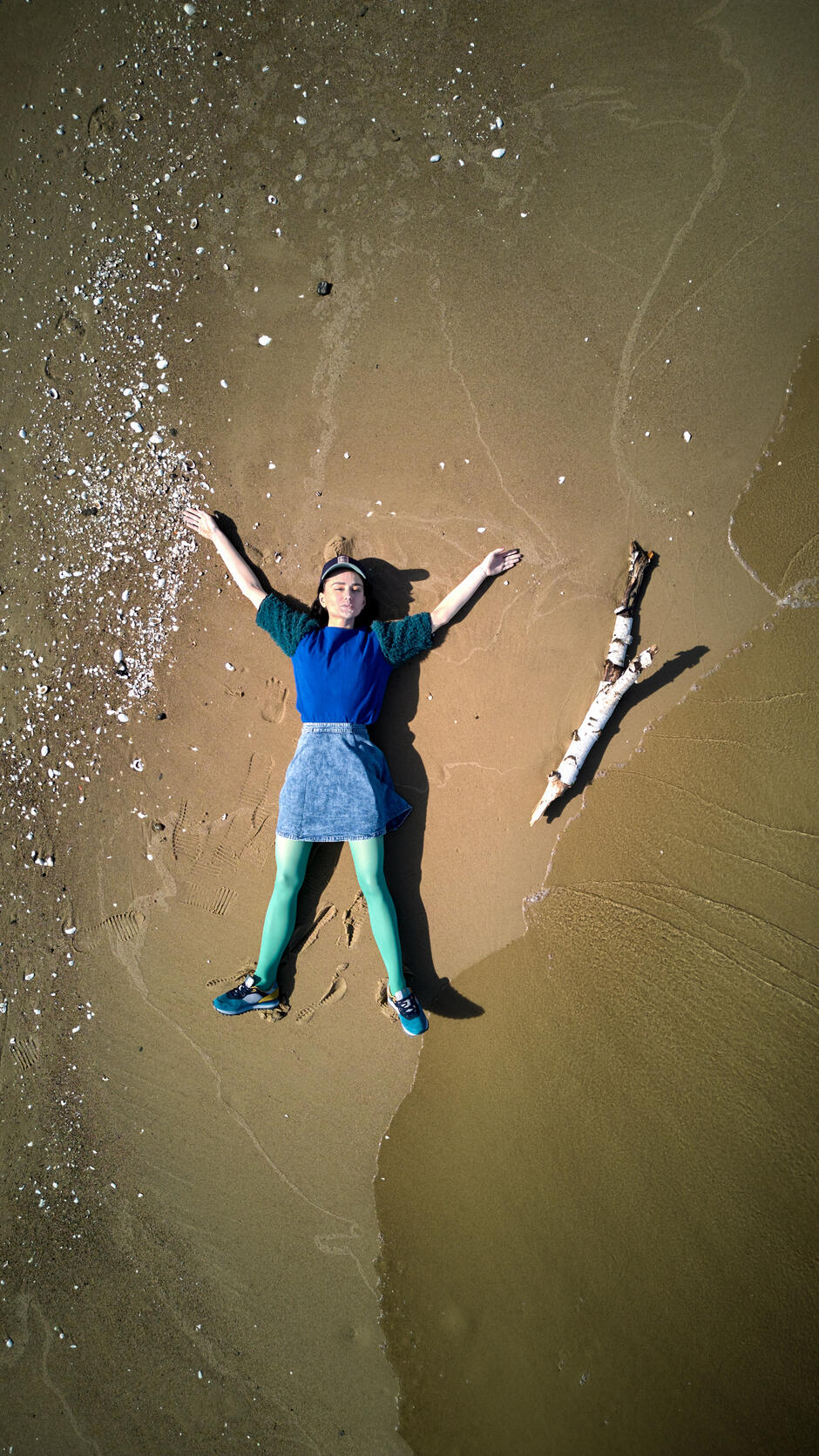 aktorka leży na piasku przy brzegu morza, obok niej duży patyk, ma ręce uniesione do góry, zdjęcie z lotu ptaka