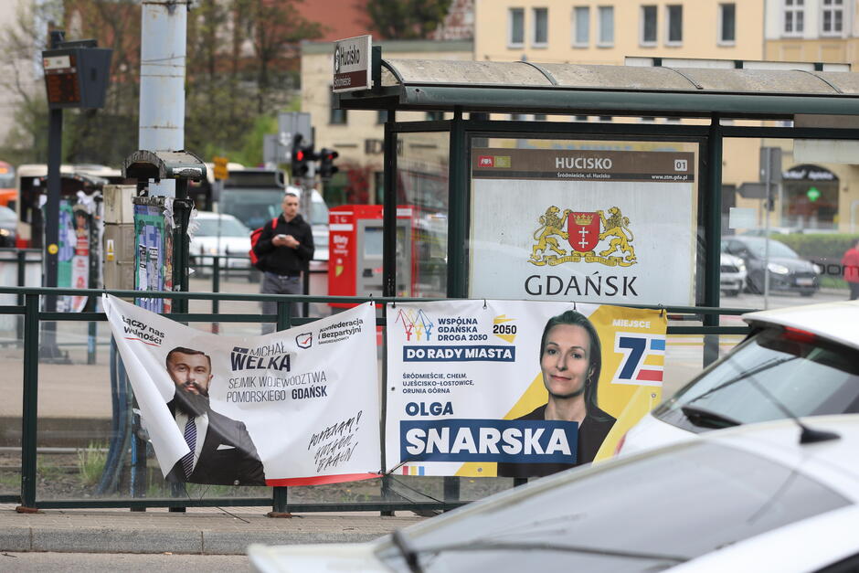 Zdjęcie pokazuje miejski przystanek autobusowy z plakatami wyborczymi umieszczonymi na jego ogrodzeniu. Na plakatach widoczne są wizerunki dwóch kandydatów, ich nazwiska oraz informacje o numerach na listach wyborczych. W tle przystanku znajduje się logo miasta Gdańsk. Przystanek jest wyposażony w siedzenia i zadaszenie, chroniące przed warunkami atmosferycznymi. W otoczeniu widać ruch uliczny, w tym przejeżdżające samochody i czerwony autobus w oddali. Na chodniku za ogrodzeniem stoi mężczyzna w czarnym stroju, co dodaje scenie miejskiego życia. Atmosfera zdjęcia jest dynamiczna i typowa dla miejskiego krajobrazu