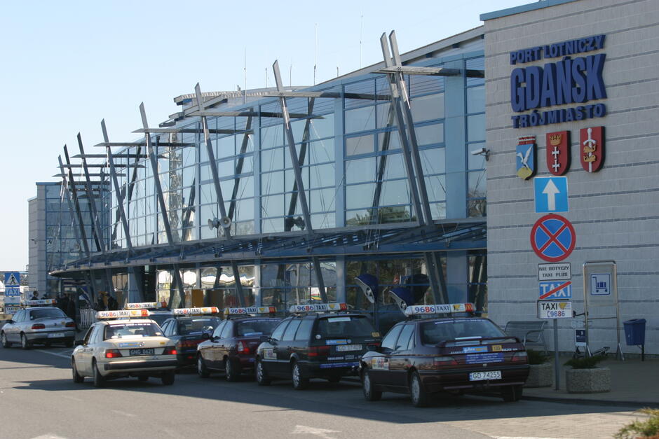 Na zdjęciu widać budynek lotniska z charakterystyczną, szklaną fasadą i wsporczymi metalowymi elementami konstrukcyjnymi. Nad wejściem widnieje napis „PORT LOTNICZY GDAŃSK TRÓJMIASTO” wraz z herbami Gdańska, Sopotu i Gdyni. Przed budynkiem stoi rząd taksówek, które wydają się być gotowe do przyjęcia pasażerów. Po lewej stronie obok taksówek znajduje się niebieski znak z białą strzałką wskazujący kierunek jazdy oraz znak zakazu zatrzymywania się z napisem „NIE DOTYCZY TAXI PLUS”. Słoneczna pogoda i cień rzucany przez pojazdy sugerują, że jest to dzień.