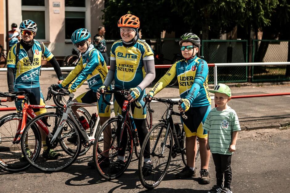 czterech rowerzystów w różnym wieku - od starszych dzieci do dorosłego, obok najmłodszy chłopiec bez roweru