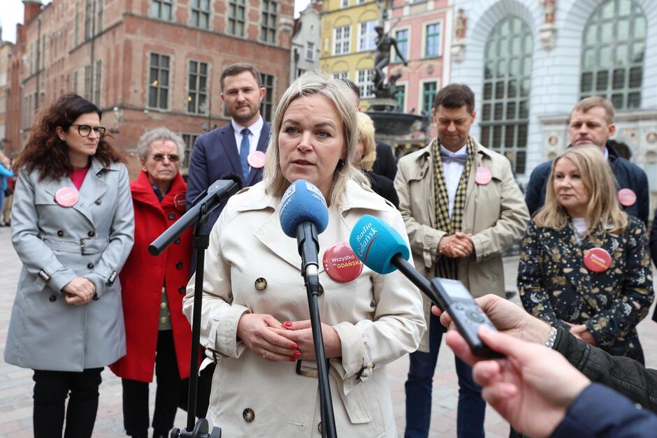 na zdjęciu kobieta w średnim wieku, w jasnym płaszczu, mówi do dziennikarskich mikrofonów, w tle za nią stoi kilka osób