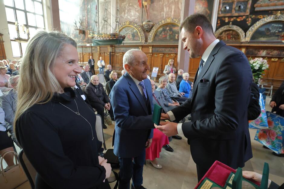 Wiceprezydent Gdańska wręcza mężczyzn starszemu mężczyźnie z okazji 60. rocznicy ślubu. Wokół na wielkiej, reprezentacyjnej sali Dworu Artusa zgromadzeni pozostali jubilaci, świętujący rocznice ślubów.