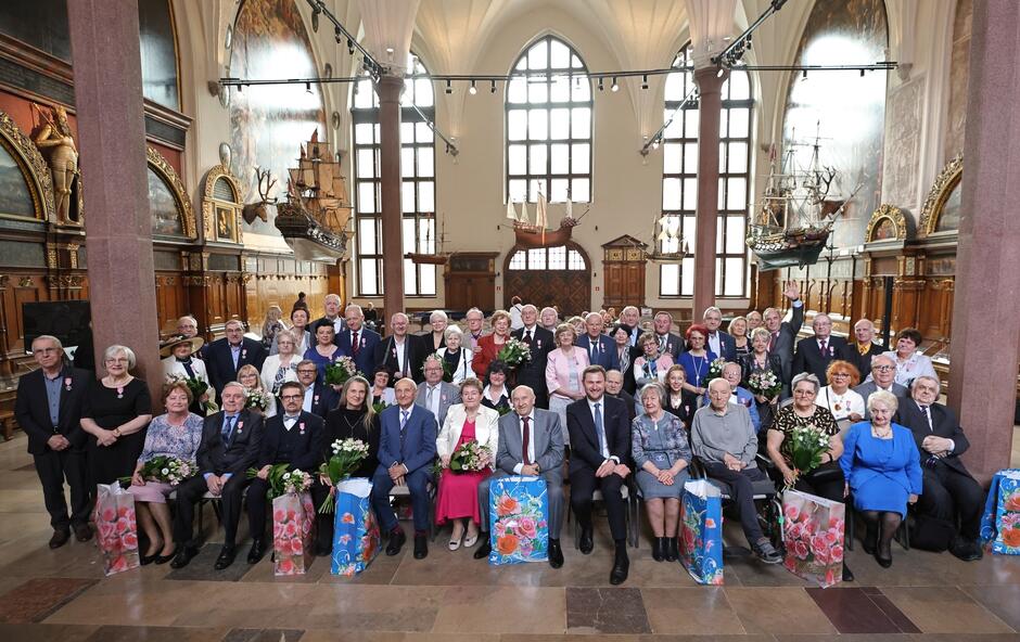 Grupa kilkudziesięciu jubilatów obchodzących złote gody oraz 55. i 60. rocznicę ślubu zebrana w wielkiej, reprezentacyjne sali Dworu Artusa.