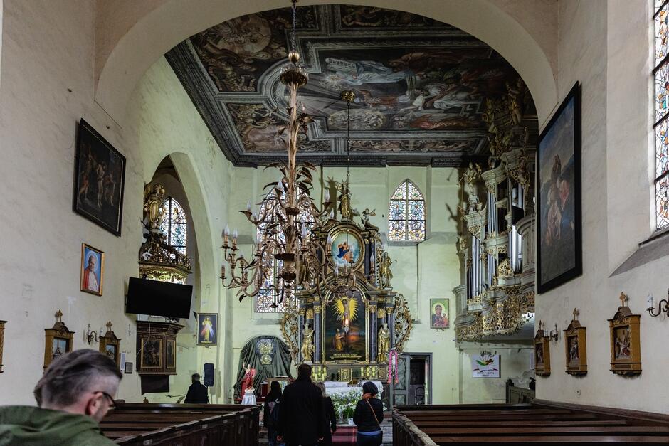 na zdjęciu wnętrze kościoła, widać ołtarz bogato zdobiony z malowidłami, widać część ławek kościelnych i kilka osób w środku