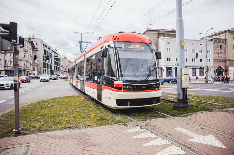 Na zdjęciu widać nowoczesny tramwaj na miejskiej ulicy. Tramwaj ma czerwono-białe malowanie z akcentami w kolorze czarnym i błękitnym, i wydaje się być stosunkowo nowym modelem. Oznaczony jest numerem 6 i dodatkowym napisem, który może wskazywać na kierunek lub przystanek docelowy. Ulica, po której porusza się tramwaj, wydaje się być częścią większego miasta z budynkami o architekturze mieszczącej się pomiędzy współczesną a tą z początku XX wieku. Widoczne są również inne pojazdy: samochody osobowe w ruchu drogowym obok tramwaju. Po prawej stronie znajduje się sygnalizacja świetlna dla pieszych oraz znaki drogowe. Pogoda wydaje się być pochmurna, a ulica jest sucha.