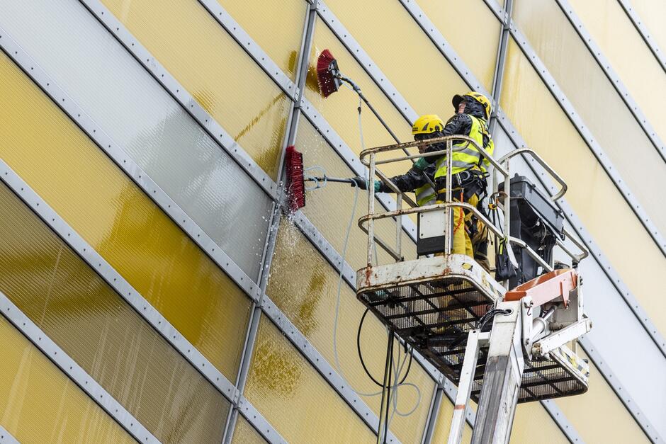 Zdjęcie przedstawia dwóch pracowników wykonujących prace na wysokości przy użyciu podnośnika koszowego. Wydają się być w trakcie czyszczenia lub malowania zewnętrznej ściany dużego budynku. Ściana składa się z pionowych paneli w odcieniach żółci i szarości. Jeden z pracowników trzyma długą szczotkę, którą aplikuje ciecz - prawdopodobnie wodę lub farbę - na ścianę, powodując spływanie płynu. Obydwaj pracownicy noszą odblaskowe kamizelki bezpieczeństwa i kaski, co sugeruje przestrzeganie zasad bezpieczeństwa. Ich postawa wskazuje na skupienie się na zadaniu.