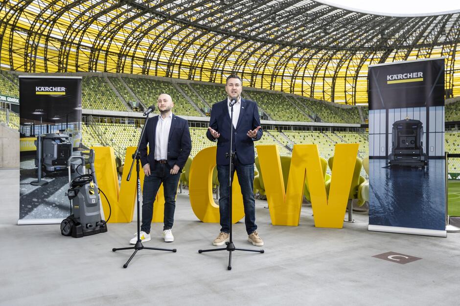 Na zdjęciu widać dwóch mężczyzn stojących przed mikrofonami wewnątrz dużego stadionu z żółtymi siedzeniami na trybunach w tle. Mężczyzna po lewej stronie stoi prosto, z rękami założonymi i z poważną miną, ubrany w jasną koszulę, ciemny garnitur i jasne buty. Mężczyzna po prawej stronie mówi i gestykuluje, jest ubrany w ciemny garnitur, białą koszulę i ciemne buty. Wokół nich znajdują się roll-upy reklamowe z marką Kärcher  i zdjęciami urządzeń czyszczących tej firmy. Na pierwszym planie po lewej stronie stoi również jedno z urządzeń. Na środku obrazu widać duże żółte litery  WOW 