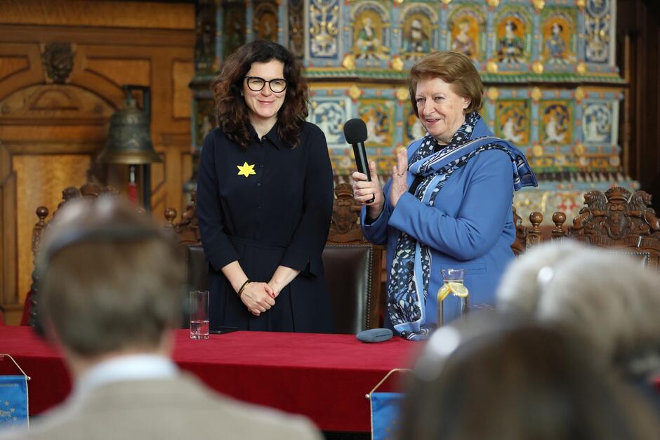 Dwie kobiety stoją za stołem w reprezentacyjnej sali Dworu Artusa, w której odbywa się międzynarodowa konferencja. Obie kobiety uśmiechają się. Jedna z nich, trzymając mikrofon bije brawo.