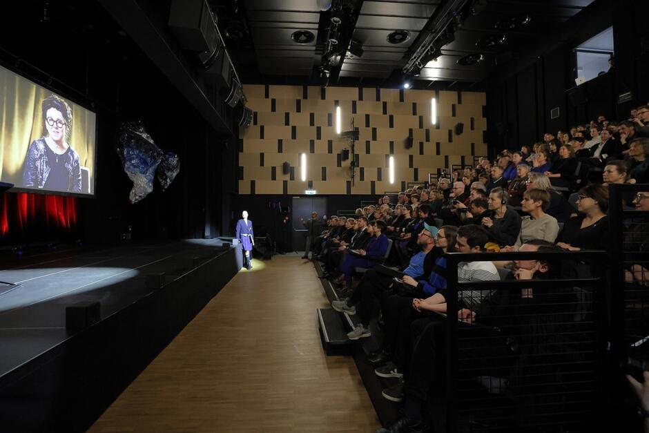 po lewej stronie ekran nad sceną, na nim wyświetlane przemówienie kobiety w okularach, z dredami, po prawej publiczność