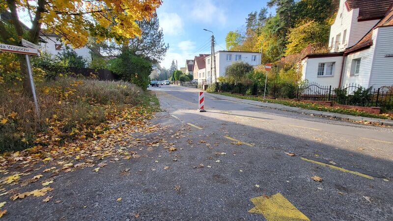 To zdjęcie przedstawia ulicę w dziennym świetle, prawdopodobnie w jesienny dzień. Widać żółte i brązowe opadłe liście, które pokrywają chodnik i krawężnik, a także żółte linie malowania drogowego. Po lewej stronie zdjęcia znajduje się znak drogowy wskazujący kierunek na Wałbrzych . Znak jest trochę przytłoczony przez gałęzie drzewa, które wpada w kadr. Po prawej stronie widoczne są domy jednorodzinne z ogrodzeniami, a dalej, wzdłuż ulicy, zaparkowane są samochody. Środek ulicy zdaje się być wolny od ruchu. Jest to spokojna, mieszkalna okolica. Światło i cienie wskazują na porę przedpołudniową lub popołudniową. Na pierwszym planie, na środku drogi, stoi pojedynczy pachołek drogowy.