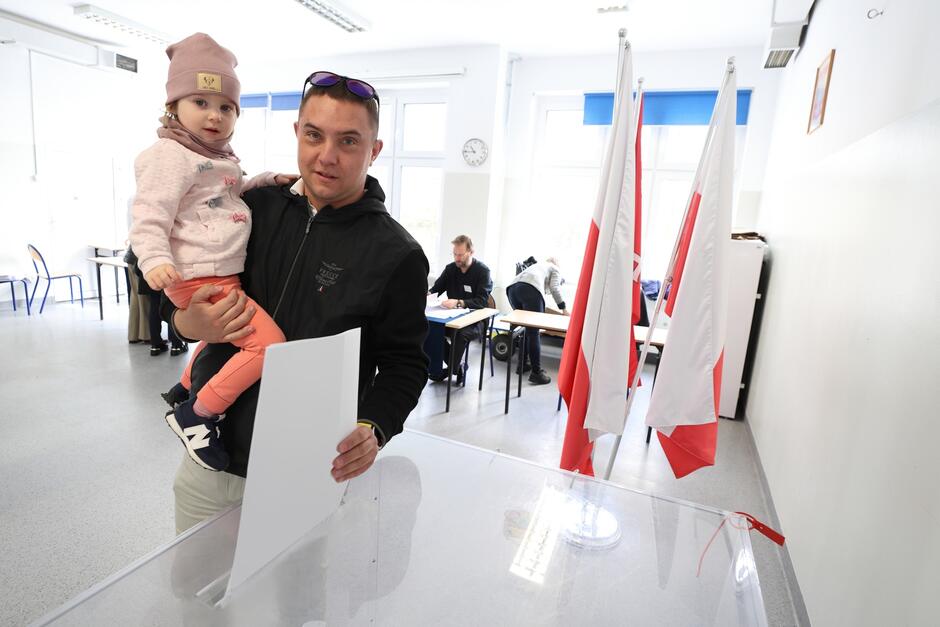 Na zdjęciu widzimy mężczyznę trzymającego na ręku małe dziecko, najprawdopodobniej jego córkę. Oboje znajdują się w jasnym pomieszczeniu z białymi ścianami, które wydaje się być lokalem wyborczym. Mężczyzna ma na sobie ciemną kurtkę i trzyma kartę do głosowania lub właśnie oddany głos, podczas gdy dziewczynka ubrana jest w różową bluzę i czapkę. W tle widoczne są polskie flagi oraz inne osoby oddające głosy za pomocą białych przegród, co sugeruje, że zdjęcie zostało zrobione podczas wyborów w Polsce. Cała scena wydaje się być pełna spokoju, a wyraźne linie prostokątnej urny i flag tworzą poczucie porządku.