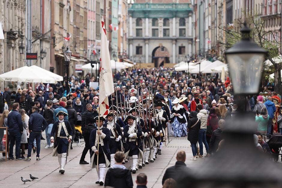 Na zdjęciu widzimy paradę odbywającą się na tle zabytkowej ulicy. Przebrani uczestnicy parady są ubrani w historyczne stroje, które przypominają XVIII-wieczne mundury wojskowe. Na pierwszym planie grupa mężczyzn niesie flagę oraz wysokie muszkiety. Uczestnicy i uczestniczki parady wydają się być częścią inscenizacji historycznej lub rekonstrukcji. Tłum gapiów po obu stronach ulicy robi zdjęcia i obserwuje przebieg wydarzenia.