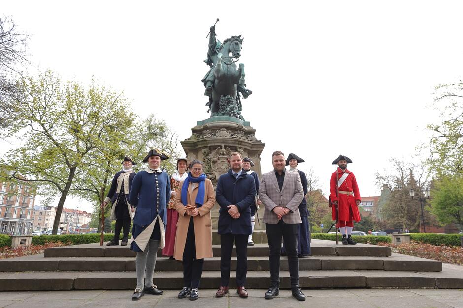 Na zdjęciu widać grupę ludzi stojących na schodach przed pomnikiem. Czterech z nich jest ubranych w stroje z epoki, które wyglądają na historyczne mundury wojskowe - dwa z lewej strony są w granatowych mundurach z epoki, a dwa z prawej strony noszą czerwone i granatowe ubiory. Pozostałe trzy osoby w środku są ubrane w współczesne ubrania. Pomnik w tle przedstawia postać na koniu, która unosi rękę, to figura związana z historią lub ważną osobistością. Sceneria wskazuje na park lub publiczne miejsce z drzewami i widoczną miejską architekturą w tle. 