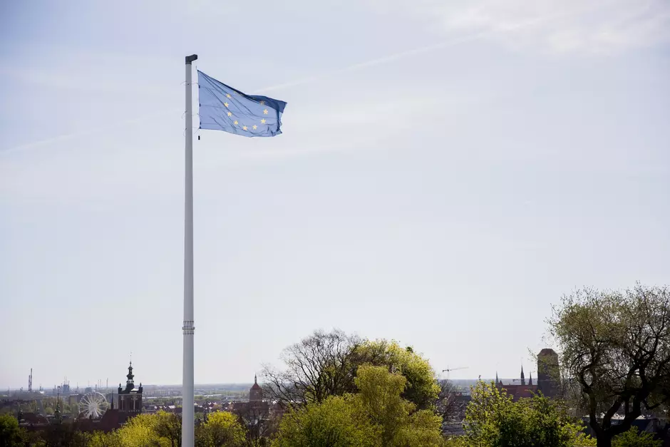 Na zdjęciu widać flagę Unii Europejskiej, która powiewa na wysokim maszcie. Flaga jest w kolorze niebieskim z kołem gwiazd w kolorze złotym, które są charakterystycznym symbolem UE. Tło obrazu to czyste, jasne niebo z kilkoma śladami kondensacji od samolotów. W dolnej części zdjęcia widoczna jest linia drzew i sylwetki budynków, co sugeruje, że flaga jest umieszczona na jakimś wzniesieniu z widokiem na miasto
