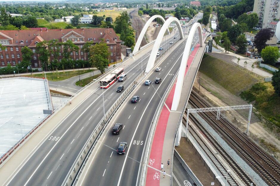 Zdjęcie przedstawia nowoczesny, miejski krajobraz z mostem nad torami kolejowymi. Most ma charakterystyczną, białą konstrukcję z trzema łukami, a jego ściany boczne są pomalowane na czerwono. Na moście wyznaczono osobne pasy: jeden dla autobusów, co wskazuje znakowanie "BUS" na drodze oraz czerwony pas, który wydaje się być przeznaczony dla rowerzystów, oznaczony także symbolami rowerów. Ruch na moście jest umiarkowany; widocznych jest kilka samochodów osobowych i autobus komunikacji miejskiej. Obok mostu, po lewej stronie zdjęcia, znajduje się duży, ceglany budynek, który może być starym budynkiem użyteczności publicznej lub mieszkalnym. Na dole zdjęcia, wzdłuż torów kolejowych, widoczna jest ścieżka dla pieszych i kilka osób spacerujących. Po prawej stronie zdjęcia, w tle, można dostrzec więcej miejskiej zabudowy. Cechą charakterystyczną tego miejsca jest współistnienie transportu publicznego, infrastruktury pieszej i kolejowej, co świadczy o dobrze zaplanowanej miejskiej przestrzeni.