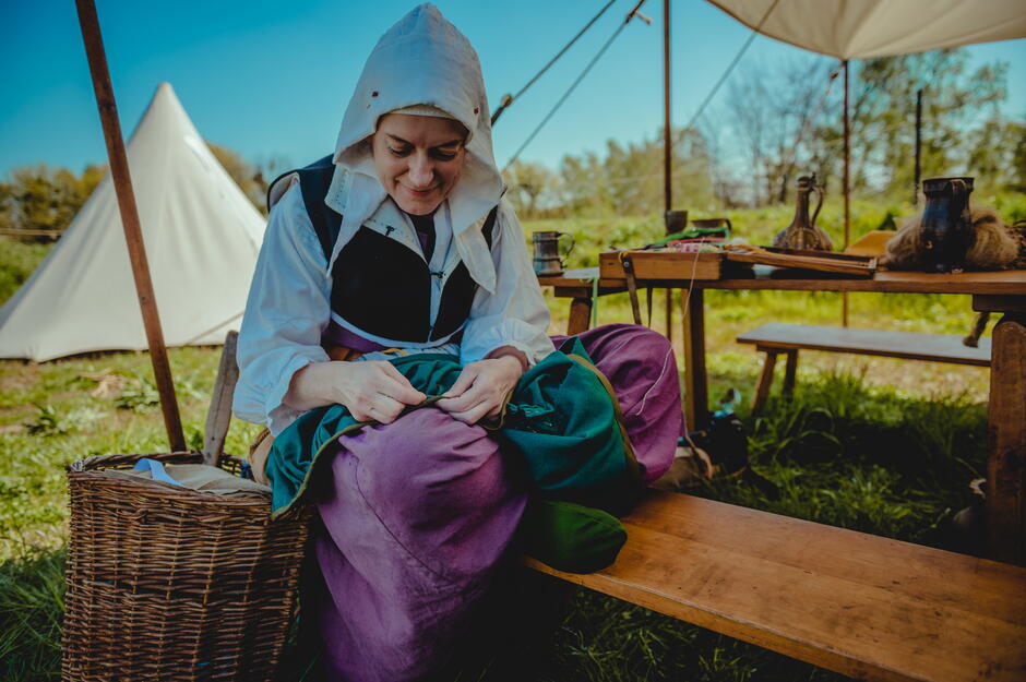 Kobieta w stroju z epoki średniowiecza siedzi na drewnianej ławce z koszem wiklinowym obok. Prowadzi pokaz krawiectwa.