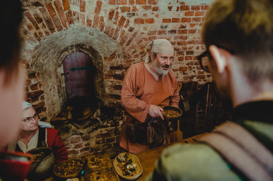 Stanowisko pokazów dawnych zawodów. Ubrany w średniowieczny strój mężczyzna z siwą brodą pokazuje miskę z różnymi przedmiotami.