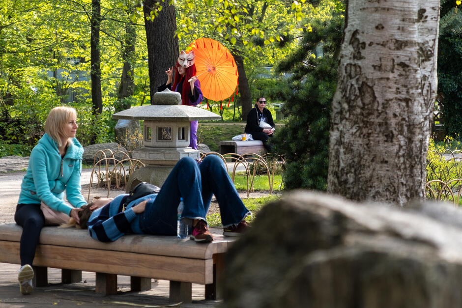 Na zdjęciu widzimy scenę w parku. Po lewej stronie kobieta w błękitnej kurtce siedzi na ławce i rozmawia z leżącą osobą, która ma na sobie ciemny sweter i dżinsy. Po prawej stronie, w tle, siedzi inna kobieta w ciemnej odzieży na innym siedzeniu, w otoczeniu zieleni i drzew. Centralnym elementem zdjęcia jest postać stojąca w środku, odziana w kolorowy, tradycyjny strój, trzymająca czerwony parasol. Na twarzy ma białą maskę z czerwonymi akcentami, która przypomina kitsune - lisa z japońskiego folkloru. Osoba ta wykonuje gest ręką, który może być interpretowany jako geście ręczne w kulturze japońskiej. Kompozycja zdjęcia, z różnymi planami i kontrastującymi elementami, tworzy ciekawy przekrój przez różne formy spędzania czasu w parku oraz połączenie kultury wschodniej i zachodniej.