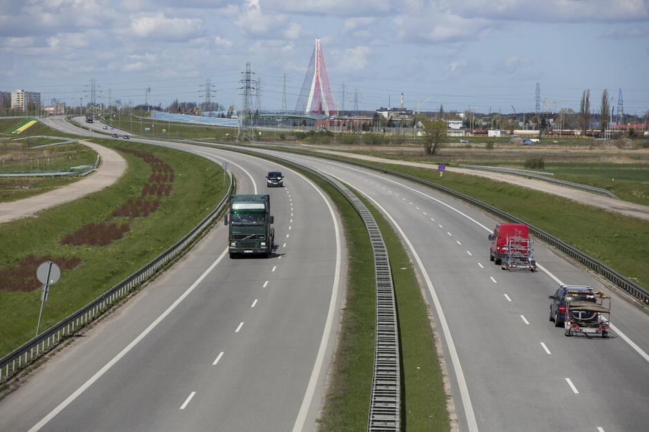 To zdjęcie pokazuje widok na autostradę w ciągu dnia. Autostrada ma cztery pasy, podzielone pasem zieleni i barierami bezpieczeństwa. Ruch jest umiarkowany, widoczne są pojazdy ciężarowe oraz osobowe. Po lewej stronie jadą dwa ciężarówki, a po prawej czerwona ciężarówka i pojazd osobowy z przyczepką. W tle dominuje unikatowy, czerwony most wiszący z białymi linami, co jest charakterystycznym elementem krajobrazu. Zdjęcie pozwala zauważyć również wysokie słupy energetyczne i niską zabudowę przemysłową w dali. Niebo jest częściowo zachmurzone, ale przebija przez nie światło słoneczne. Teren wokół autostrady jest otwarty, z niewielką ilością zieleni.