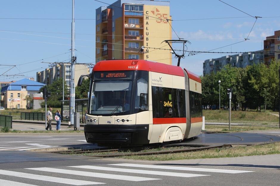 Na zdjęciu widoczny jest czerwono-biały tramwaj na miejskim torowisku, oznaczony numerem 11 i napisem CHEŁM HITOSA PRZEZ BRZESZCZ . Model tramwaju to Swing, co jest widoczne po napisie na boku. Tramwaj znajduje się w ruchu, zwrócony w lewą stronę zdjęcia. W tle widzimy miejski krajobraz, w tym bloki mieszkalne o różnej architekturze i kolorach, a także zielone tereny z drzewami. Na chodniku obok torów widoczne są dwie osoby, prawdopodobnie pasażerowie lub przechodnie. Jest to jasny, słoneczny dzień, a na niebie brak chmur. Atmosfera wydaje się być spokojna, typowa dla miejskiego krajobrazu w ciągu dnia