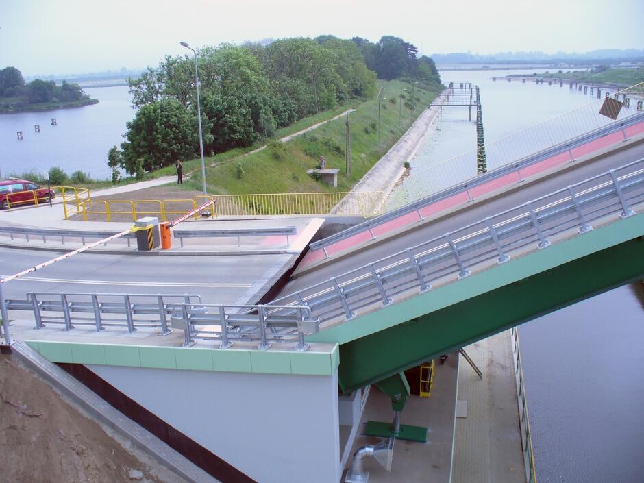 To zdjęcie pokazuje nowoczesną konstrukcję mostu zwodzonego. Most ma metalową konstrukcję, pomalowany jest na jasnoszaro z zielonymi elementami. Wyposażony jest w balustrady i barierki bezpieczeństwa. Prowadzi nad kanałem, który łączy dwie większe zbiorniki wodne. Z prawej strony mostu widać drogę dojazdową i fragment nabrzeża, a także zarośnięty brzeg przeciwległy. Na dole, po lewej stronie, znajduje się żółta zapora drogowa, a za nią widać czerwony samochód oraz kilka drzew. Pogoda wydaje się być pochmurna, co wskazuje na chłodniejszy klimat lub porę roku.