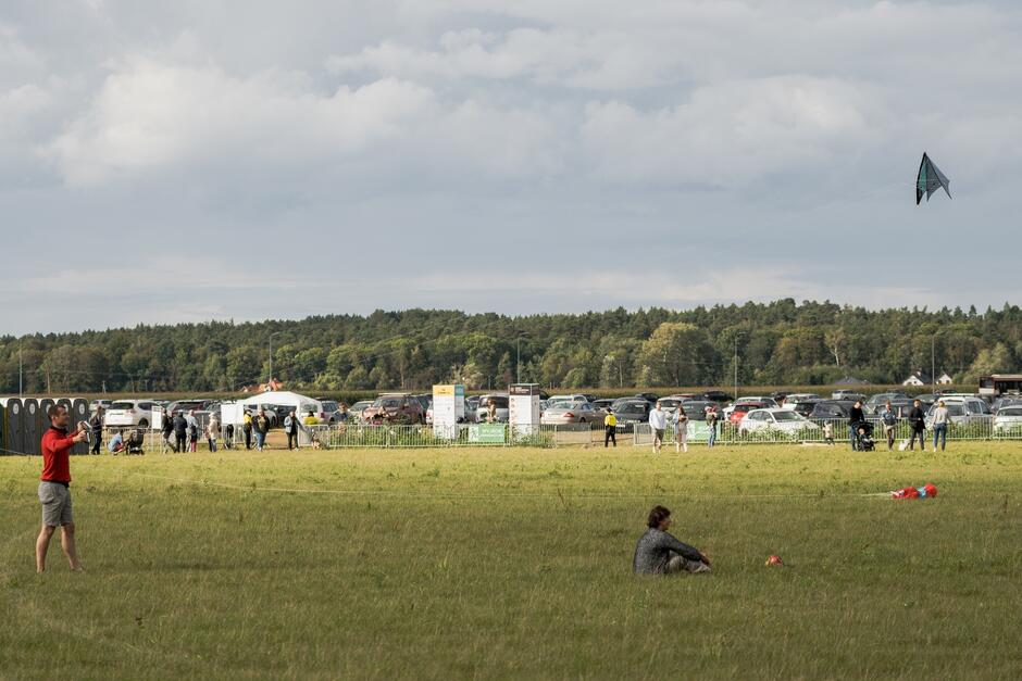 Zdjęcie przedstawia rozległą, trawiastą przestrzeń, na której ludzie bawią się na zewnątrz. W centralnej części zdjęcia widać mężczyznę w czerwonych szortach, który właśnie puszcza latawiec. Niebo jest lekko zachmurzone, co sugeruje przyjemny, umiarkowany dzień. W tle można dostrzec lasy oraz parking z wieloma samochodami, co wskazuje na to, że miejsce to jest popularnym punktem spotkań lub odbywa się tutaj jakiś event. Cała scena oddaje atmosferę relaksu i rekreacji na świeżym powietrzu