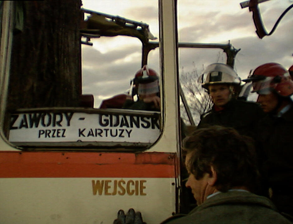 Fragment rozbitego autobusu na drzewie. Na drzwiach widać tabliczkę z kursem: Zawory - Kartuzy - Gdańsk. Obok stoją strażacy.