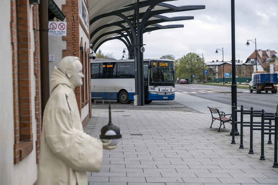 Pod wiatą autobusową na przystanku stoi autobus. Na pierwszym planie budynek dworca autobusowego i rzeźba mnicha.