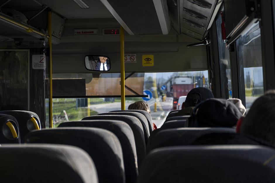 Wnętrze autobusu, na niektórych miejscach siedzą pasażerowie, w lusterku widoczne odbicie kierowcy, przez przednią szybę widać korek na szosie.