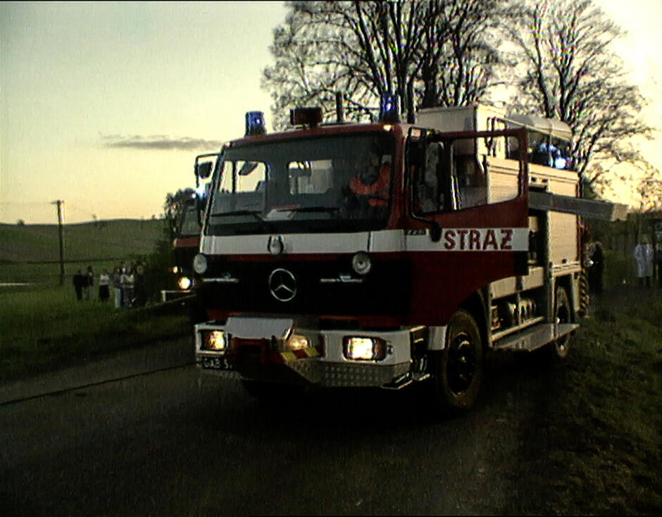 Wóz strażacki marki mercedes migający kogutami stoi na szosie. W tle przypatrują się ludzie.