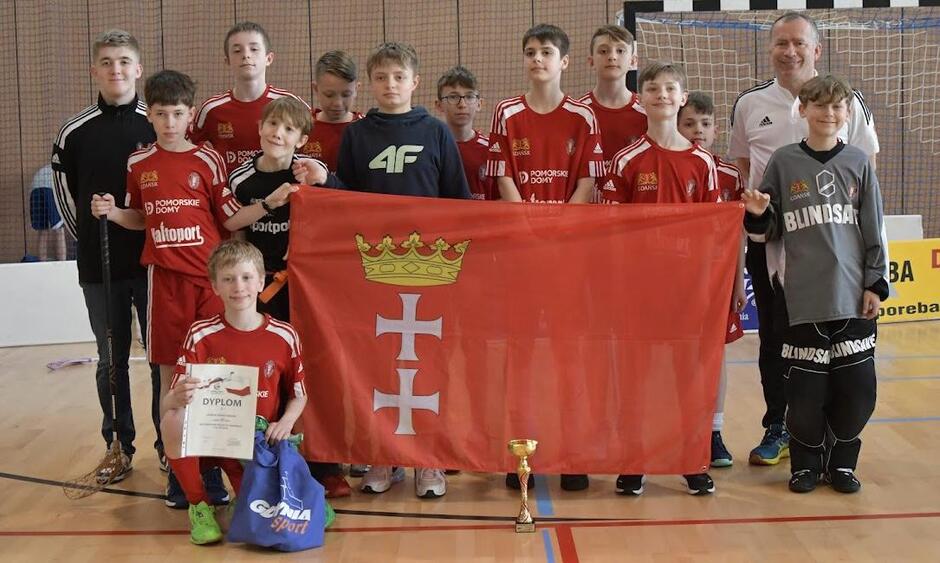 Chłopcy na sali sportowej w strojach i dresach. Obok troi po prawej trener. Chłopcy trzymają czerwoną flagę z godłem Gdańska. Jeden klęczy i trzyma w dłoni dyplom