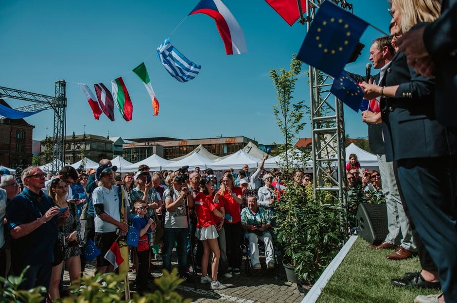 Zdjęcie przedstawia zgromadzenie ludzi na zewnętrznym wydarzeniu, prawdopodobnie festiwalu lub publicznym zgromadzeniu, które ma miejsce w słoneczny dzień. Widoczni uczestnicy, zróżnicowani pod względem wieku, skupiają swoją uwagę na mówcach stojących na scenie. Scena jest otoczona przez flagi różnych krajów, w tym flagi narodowe i flagi Unii Europejskiej.
Na scenie znajdują się dwie postacie, które aktywnie przemawiają do publiczności, tłum reaguje, niektórzy z uczestników trzymają małe flagi, co dodatkowo podkreśla celebracyjny i oficjalny charakter zgromadzenia.
W tle można dostrzec namioty. 